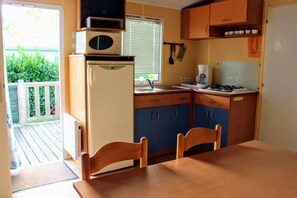 A compact kitchen with a refrigerator, microwave, and stove. There is a window with blinds, a sink, and a wooden table with chairs. - ZEcamping 3* piscine chiens acceptés locatif TV BBQ 32m2 6 pers (Verteillac)
