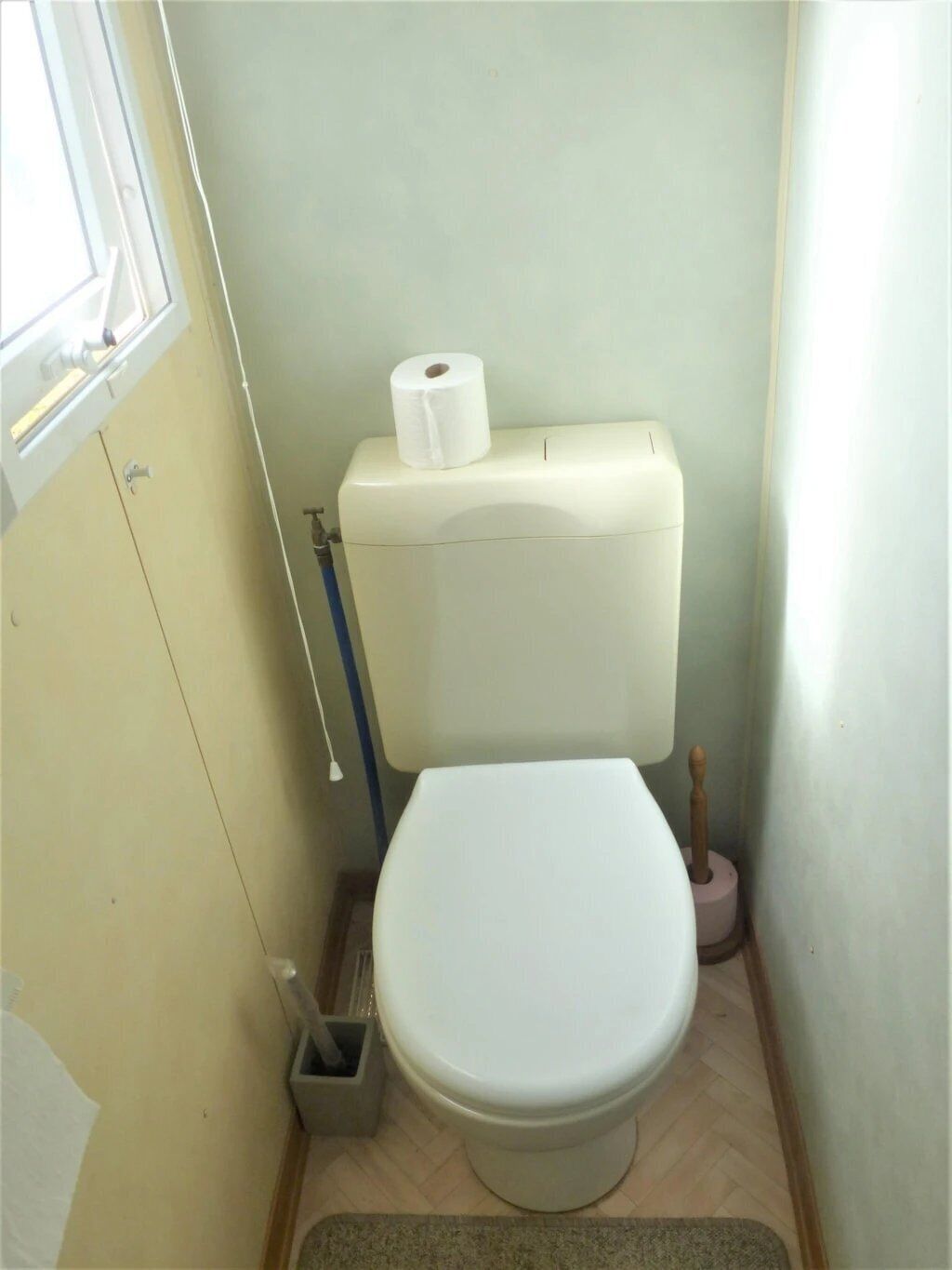 A small, compact bathroom with a white toilet, a roll of toilet paper on the tank, a bidet, and a wooden bidet brush.