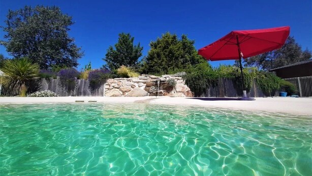Piscina cristalina con una estructura de piedra al fondo, rodeada de árboles y una sombrilla roja.