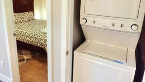 Een compacte wasmachine en droger in een slaapkamer, met een bed met een geborduurde spreideken en een houten hoofdeinde.