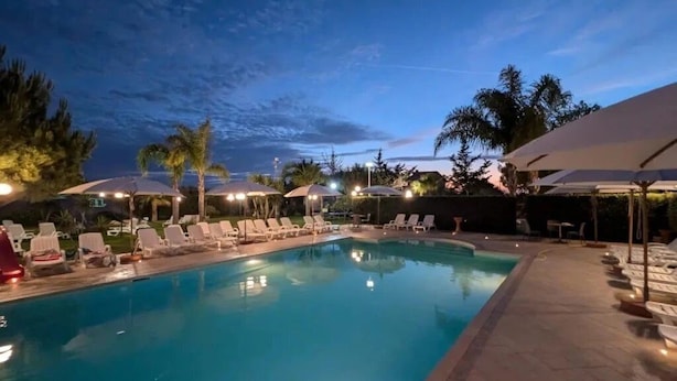 Ein Swimmingpool mit Liegestühlen und Sonnenschirmen, umgeben von Palmen und blauem Himmel.