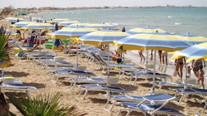 Ein Strand mit Reihen von Liegestühlen und Sonnenschirmen, ein sandiger Uferstreifen und das Meer im Hintergrund.