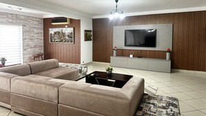 Een moderne woonkamer met een grote flatscreen televisie aan een houten wand, een beige bank, een salontafel en een klein bijzettafeltje met decoratieve objecten.