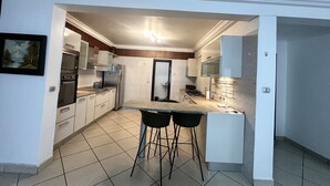 Een kleine keuken met witte kastjes, een ingebouwde oven, een magnetron en een koelkast. Er zijn twee zwarte barkrukken en een klein aanrecht met een spoelbak.