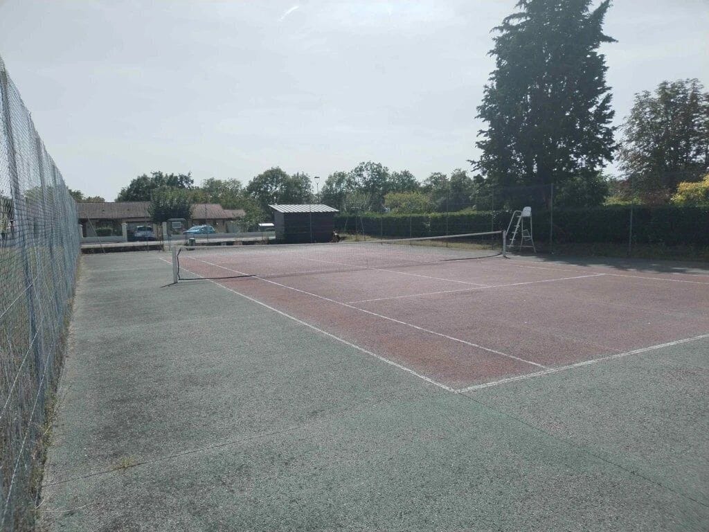 Een buitenbaan voor tennis met een net, omringd door een hek, en een klein gebouw op de achtergrond.