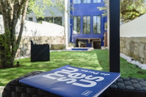 Un livre intitulé « mundo de portos » est posé sur une chaise en osier dans un jardin, avec un bâtiment bleu en arrière-plan.