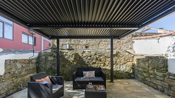 An outdoor patio with a covered seating area, stone walls, and a view of a building in the background. - GuestReady - Alegria 293 - Room 1 (Porto)
