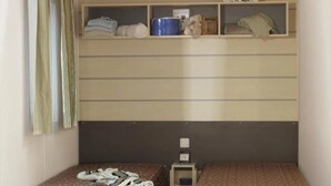 Ein kleines Zimmer mit zwei Einzelbetten, einem Regal mit persönlichen Gegenständen und einem Fenster mit Vorhängen.
