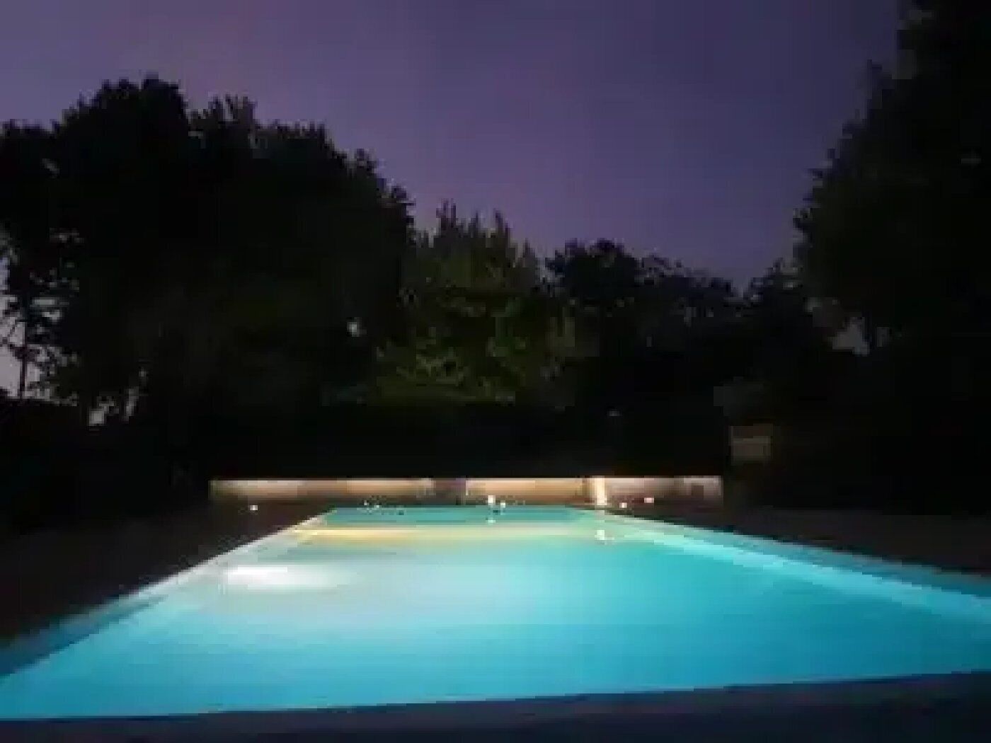 Ein rechteckiger Swimmingpool mit beleuchtetem Rand, umgeben von Bäumen und einem dunklen Himmel.