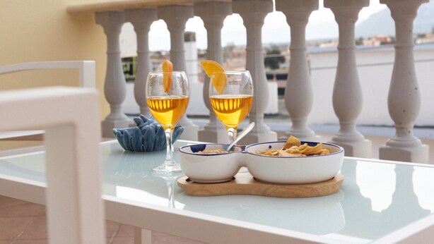 A table with two glasses of a golden beverage, a bowl with chips, and a blue bowl with a spoon on a balcony. - Casa Duesse (IUN R6224)Accommodation located in the very center of Orosei. (Orosei)