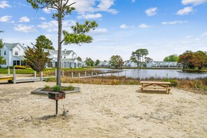 Une plage de sable avec un banc en bois et un petit arbre, une étendue d’eau, et une rangée de maisons blanches en arrière-plan.