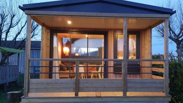 A wooden cabin with a covered porch, large windows, and a dining area visible inside. - ZEcamping 3 étoiles piscine chiens acceptés locatif TV 28m2 6 pers (La Tremblade)