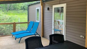 A covered outdoor area with a wooden deck, two blue lounge chairs, a table, and a door leading to a glass door.