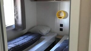 Ein kleines Zimmer mit zwei Etagenbetten, einem kleinen Fenster mit Vorhang und einem gelben Wandregal.