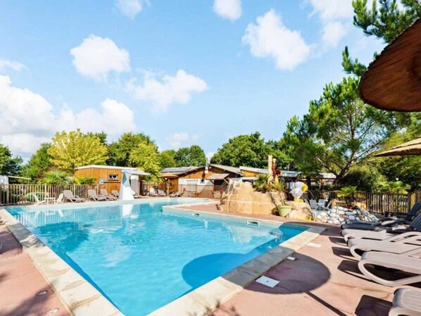 Piscina com espreguiçadeiras, toboágua e cabanas de madeira ao fundo.