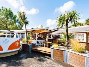 Uma carrinha VW camper estacionada numa área exterior soalheira com um alpendre de madeira, mobiliário de exterior e plantas tropicais.