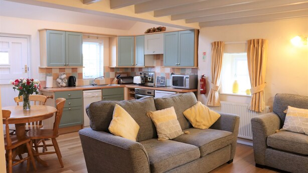 Una sala de estar acogedora con un sofá gris, una mesa de comedor de madera y sillas. Se ve una cocina con armarios color turquesa y electrodomésticos modernos al fondo.