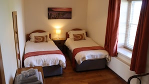 A room with two beds, a bedside table with a lamp, a window with curtains, and a painting on the wall.