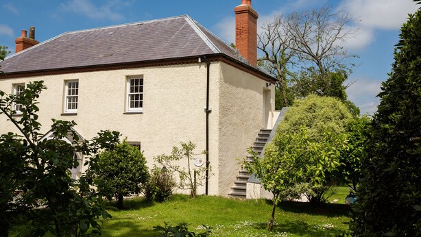 Une maison à deux étages, dotée d’un toit en pente, d’une cheminée en briques et d’un petit jardin à l’avant, avec un chemin menant à l’entrée.