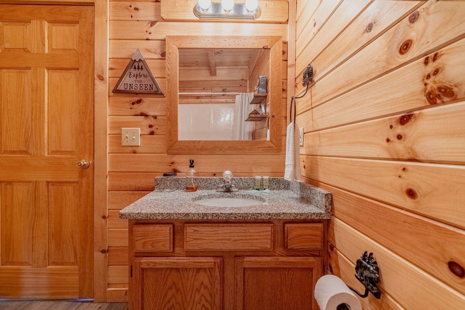 A wooden cabin bathroom with a granite sink, wooden cabinets, a towel rack, and a sign that says "Explore the Unseen."