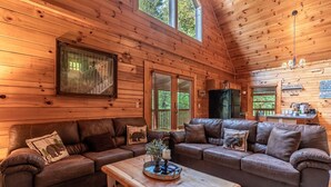 Interior acogedor de una cabaña con un amplio sofá de madera, una mesa de centro de madera y una zona de cocina con un refrigerador y un letrero que dice 'Lake Run Cabin'.