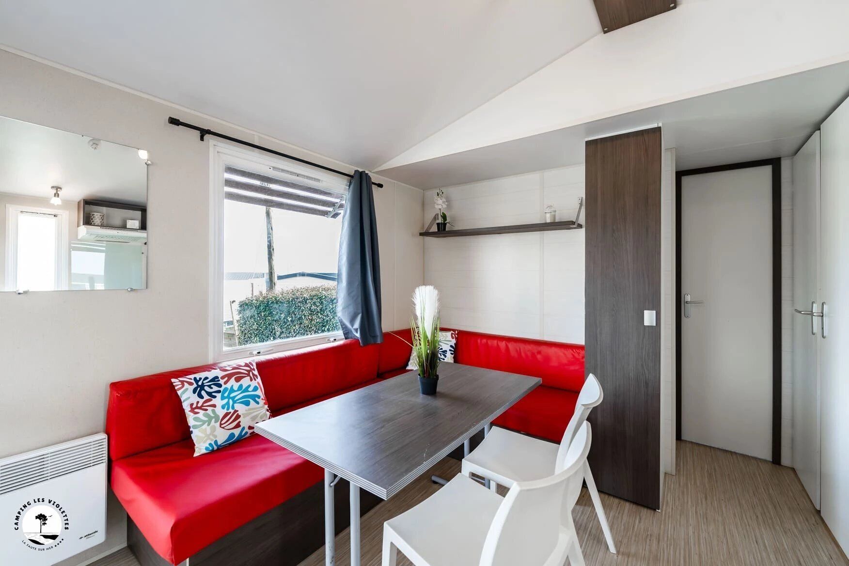 A compact living space with a red sofa, a dining table, and white chairs. A window with a view of greenery is visible, and a mirror hangs on the wall.