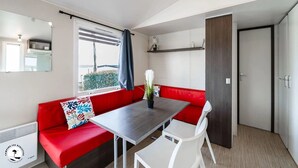 A compact living space with a red sofa, a dining table, and white chairs. A window with a view of greenery is visible, and a mirror hangs on the wall.