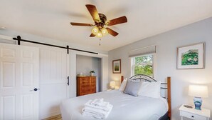 Ein Schlafzimmer mit einem Bett, einem Deckenventilator, einem Nachttisch, einer Kommode, einem Fenster und einer Tür.
