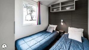 Een slaapkamer met twee bedden, een raam met gordijnen, een plank boven de bedden en een wandlamp.