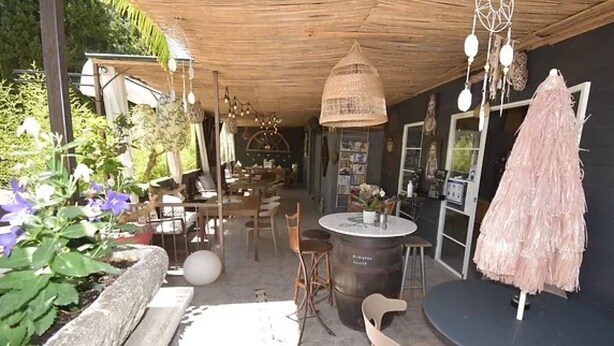 Un patio al aire libre con una mesa redonda, sillas y una hamaca colgante. Hay plantas y un paraguas decorativo.