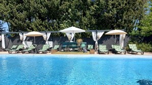 Zona de piscina con sillones de descanso y sombrillas, rodeada de árboles.