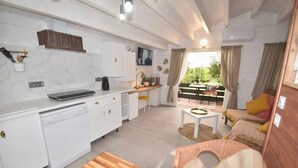 Cocina compacta con armarios blancos, una cafetera y un televisor. Hay un comedor con mesa y sillas, y una pequeña zona de estar con cojines.