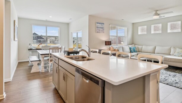 A modern kitchen with a central island, a dining area with a table and chairs, a sofa, and a view of houses through large windows. - Beach Club Getaway! Ample Amenities and Short Drive to Beaches! (OceanView)