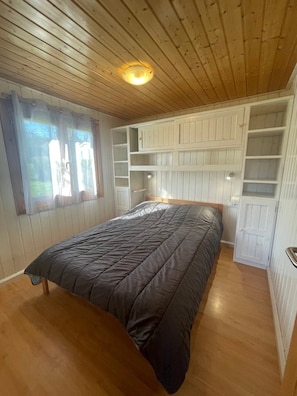 A bedroom with a bed, wooden ceiling, built-in shelving, and a window with curtains.