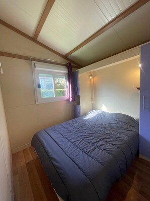 A single bed with a blue cover in a room with a sloped ceiling, a window with curtains, and a wall-mounted light. - Chalet 3 étoiles - Piscine  - eehgfd (Castelnau-Picampeau)