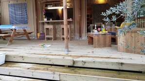 Une terrasse en bois rustique, avec une table et des chaises en bois, un luminaire suspendu et une structure en bois dotée d’une fenêtre.