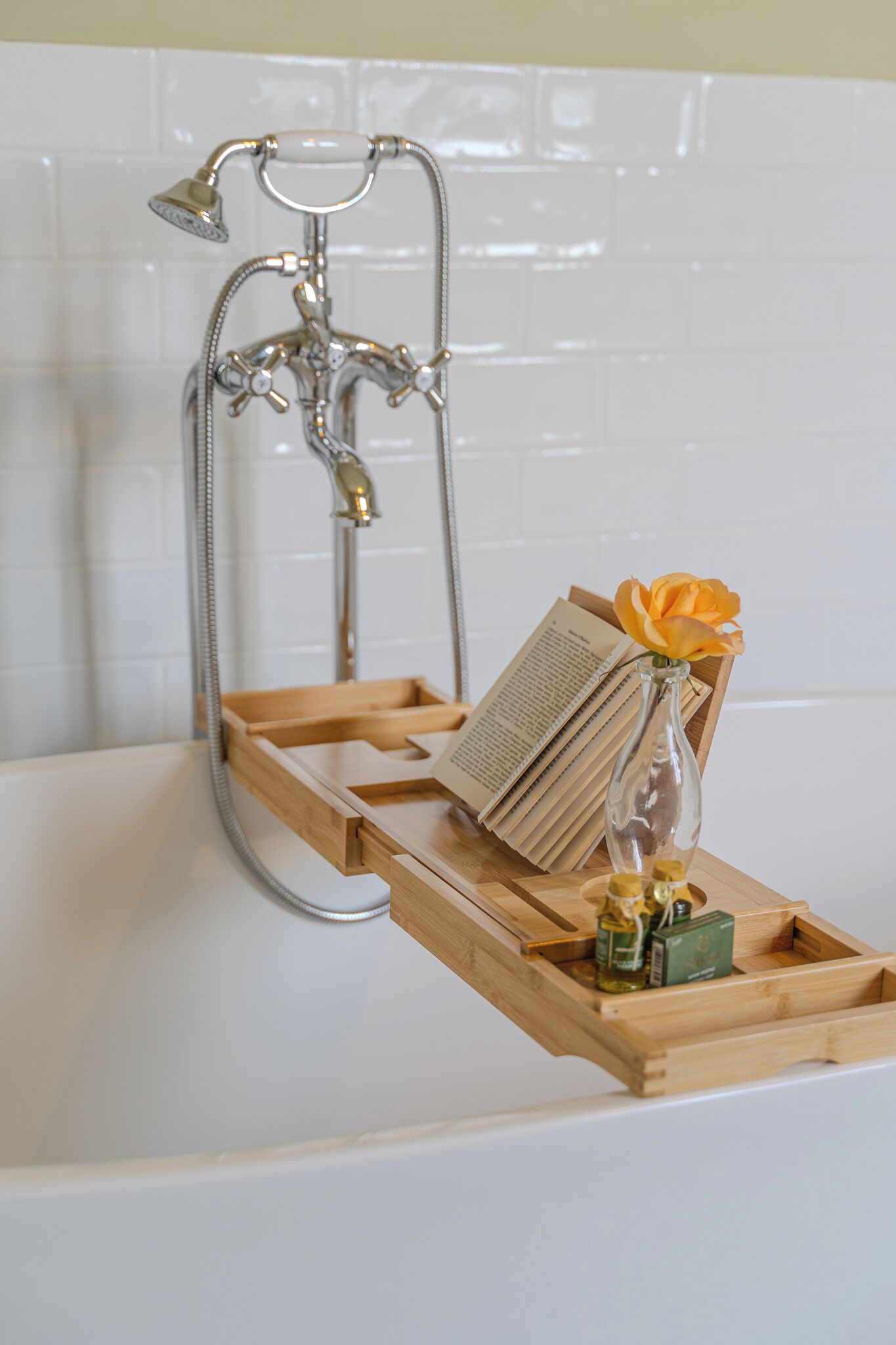 Un baño con una bañera, una bandeja de madera, un libro y un florero con una flor.