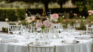 Una mesa redonda dispuesta para un evento formal, con manteles blancos, cristalería transparente y flores rosas.