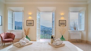 Un dormitorio amplio con una cama grande, dos ventanas que ofrecen vistas a un paisaje montañoso, una silla rosa y jarrones decorativos.
