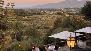 Un área de comedor al aire libre con mesas y sillas, rodeada de grava, con vistas a un paisaje pintoresco con montañas y un pueblo a lo lejos.
