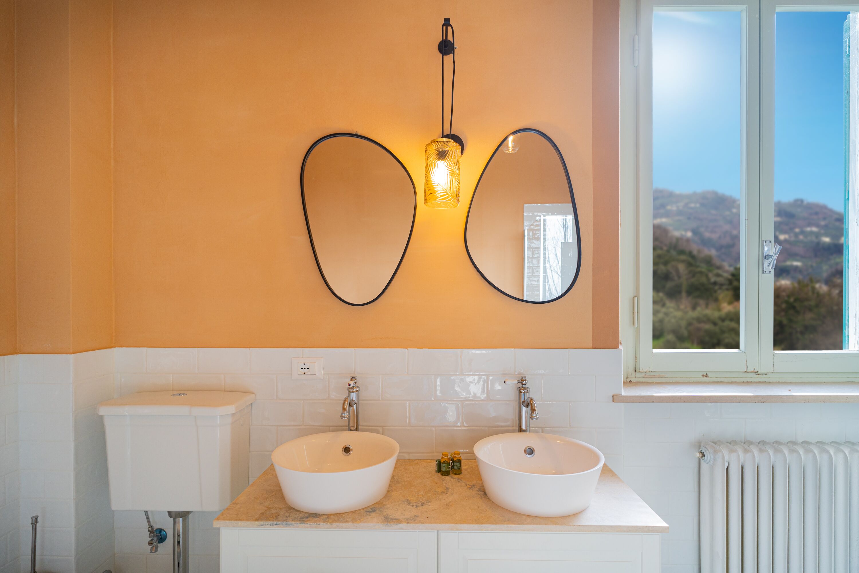 Une salle de bain avec deux lavabos, des toilettes et une fenêtre donnant sur des collines.
