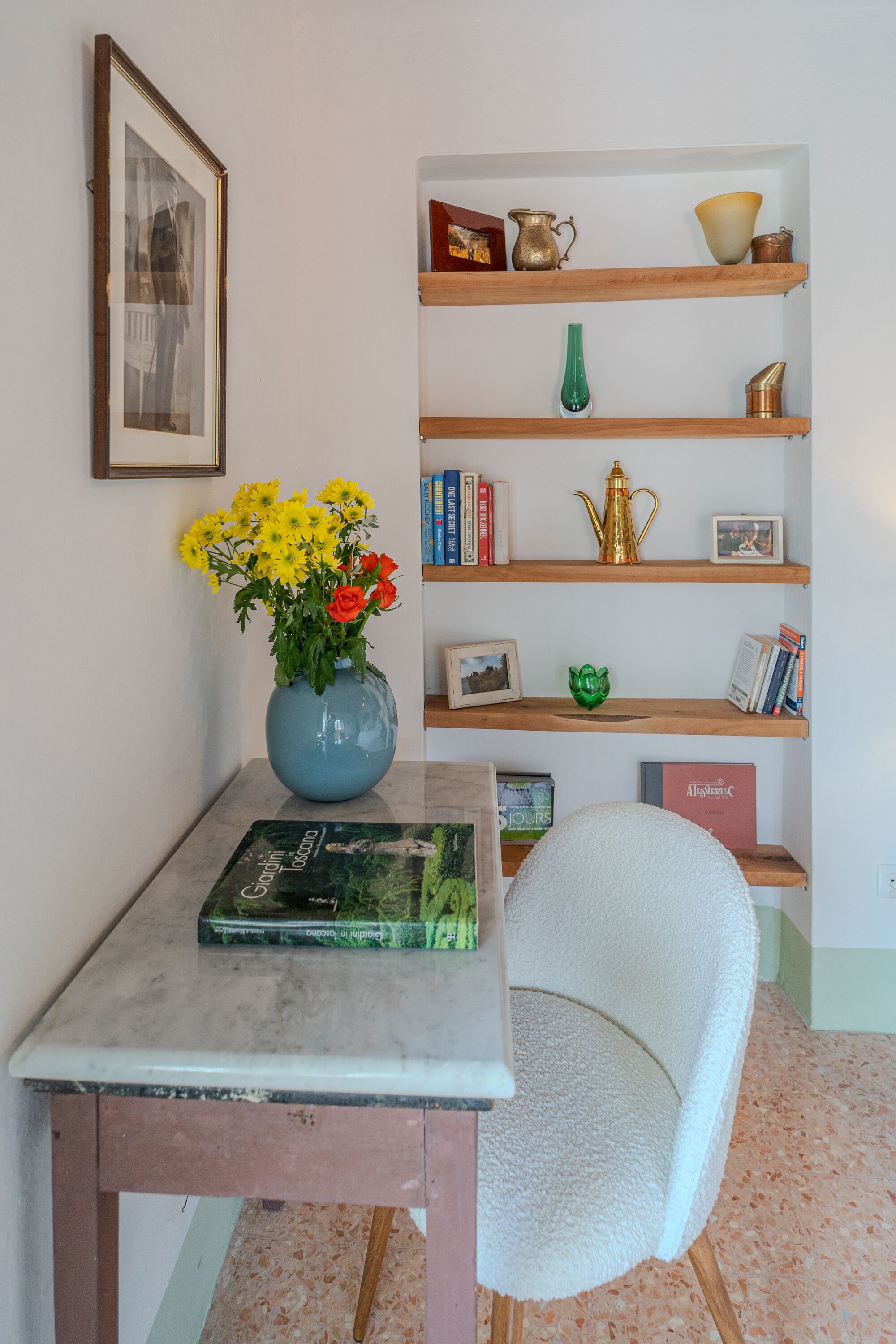 Una stanza con un tavolo in marmo, un vaso blu con fiori, una sedia bianca e mensole in legno con libri e oggetti decorativi.