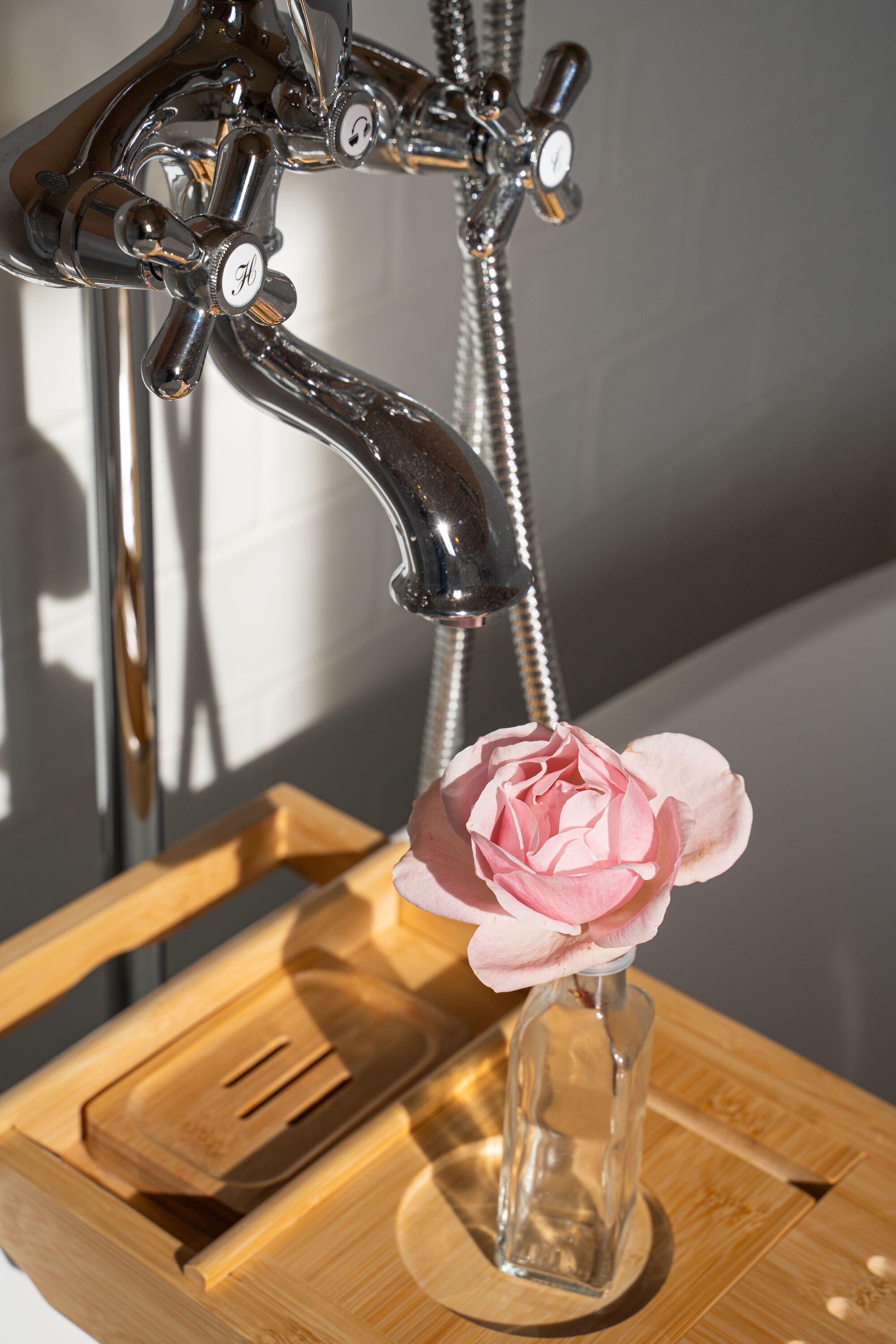 A bathroom with a chrome faucet, a shower head, and a wooden tray holding a glass vase with a pink flower.