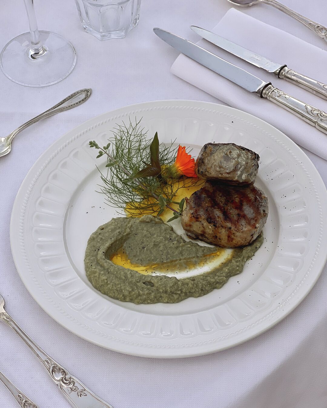 Ein weißer Teller mit gegrilltem Fleisch, grüner Soße und einer Garnitur auf einem Tisch mit Besteck und einem Glas.