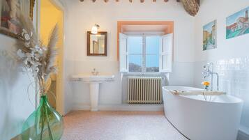 Un baño con una bañera independiente, un lavamanos sobre pedestal y una ventana con contraventanas abiertas.