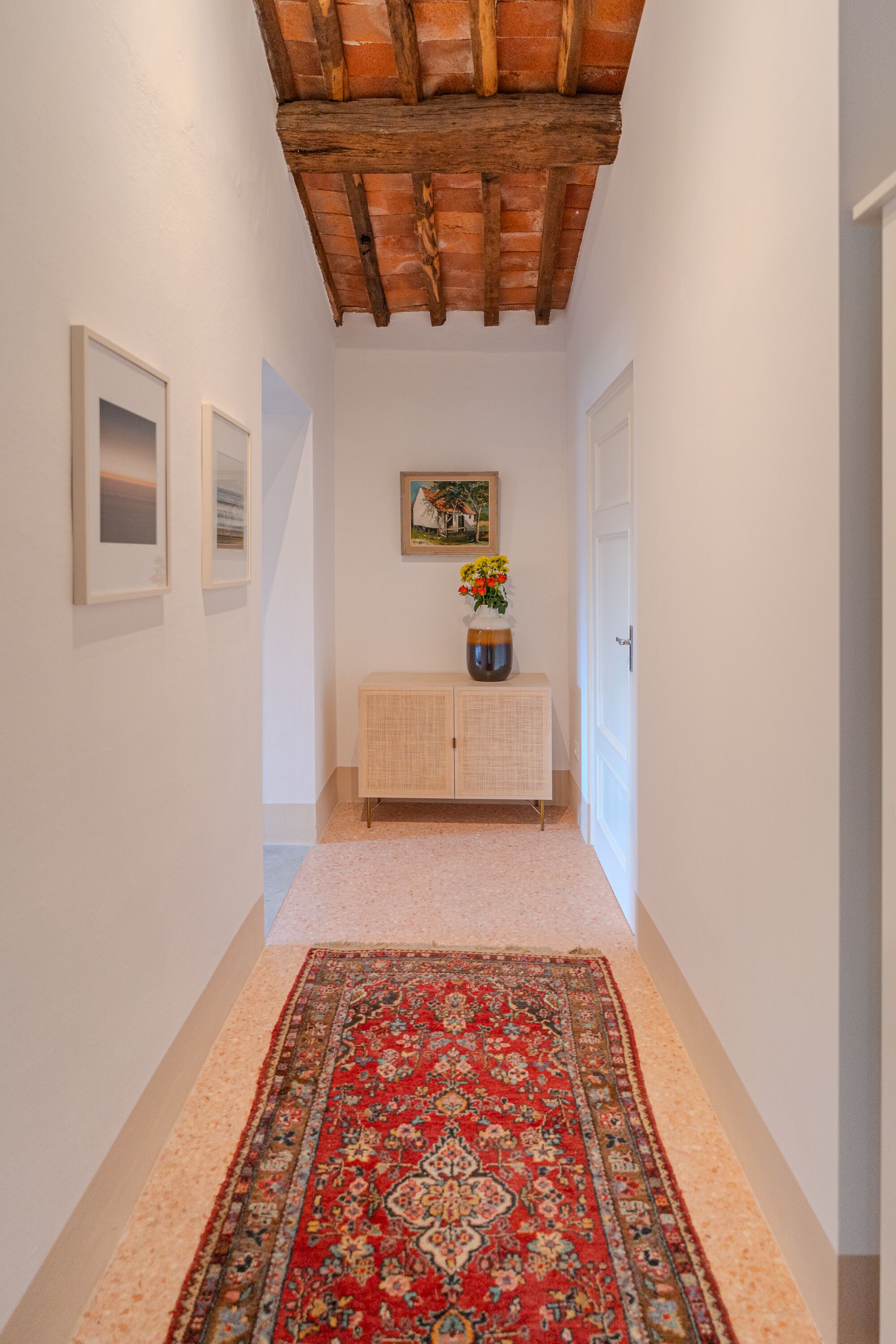 Un pasillo con un tapete rojo estampado, techo de madera, una puerta blanca y un armario con un jarrón de flores.