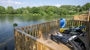 A wooden deck with lounge chairs and a barbecue grill overlooking a calm lake surrounded by dense greenery.