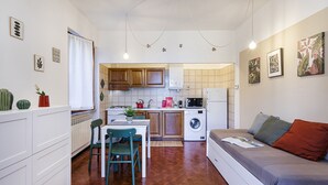 A compact kitchen with wooden cabinets, a dining area with a table and chairs, a washing machine, and a sofa with cushions. - Una Palma a Camogli by Wonderful Italy (Camogli)