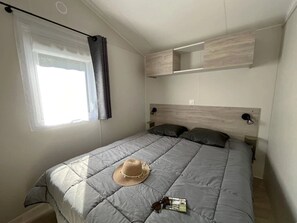 Ein ordentlich gemachtes Bett mit einer grauen Decke, einem Strohhut und Sonnenbrille darauf. Ein Fenster mit Vorhang, ein Regal über dem Bett und eine an der Wand befestigte Lampe sind ebenfalls zu sehen.