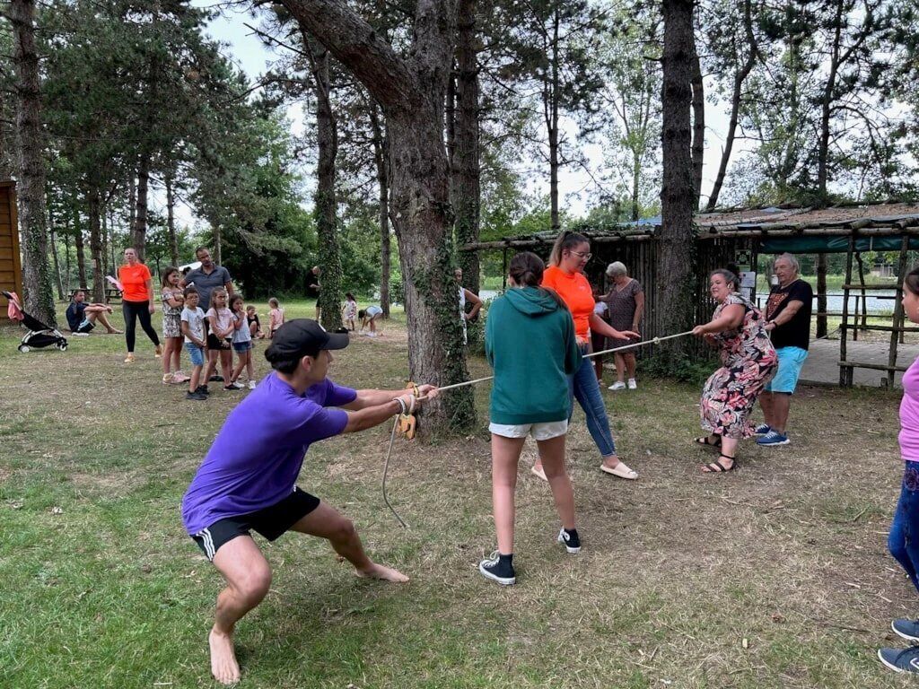 Un gruppo di persone che giocano a tirare la corda in un ambiente esterno, con alberi e una struttura in legno sullo sfondo.